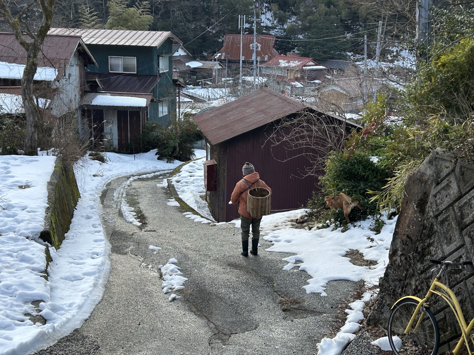 雪の残る集落を籠を背負って歩く